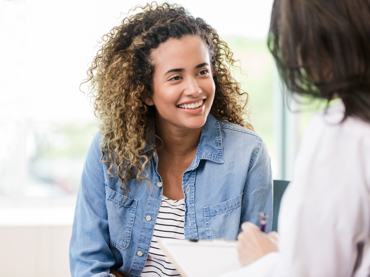 A smiling young woman with curly hair sits across from a healthcare provider during a consultation. The provider is holding a clipboard and pen, and the woman looks engaged and comfortable in the conversation.