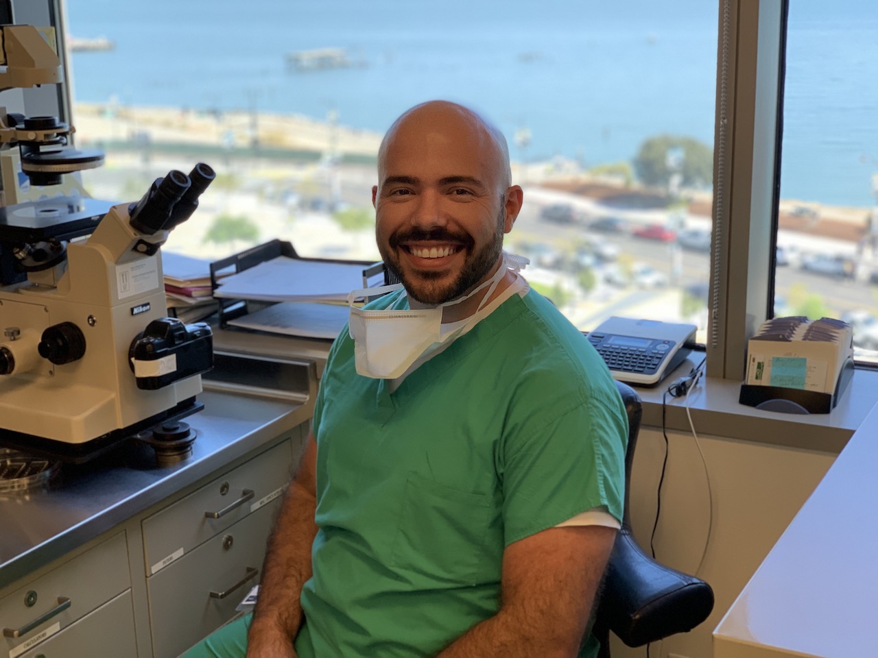 Salustiano smiling at the camera while at work with the San Francisco Bay in the window behind him